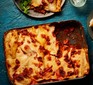 Baking dish of lasagne with a portion removed and put on plate with a green salad